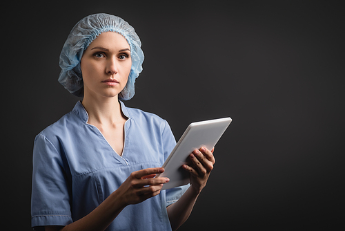 nurse in medical cap and uniform holding digital tablet isolated on dark grey