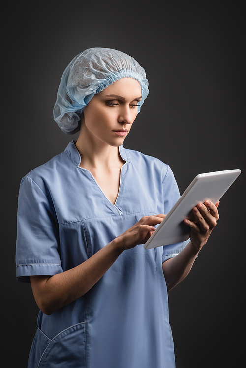 nurse in medical cap and uniform using digital tablet isolated on dark grey
