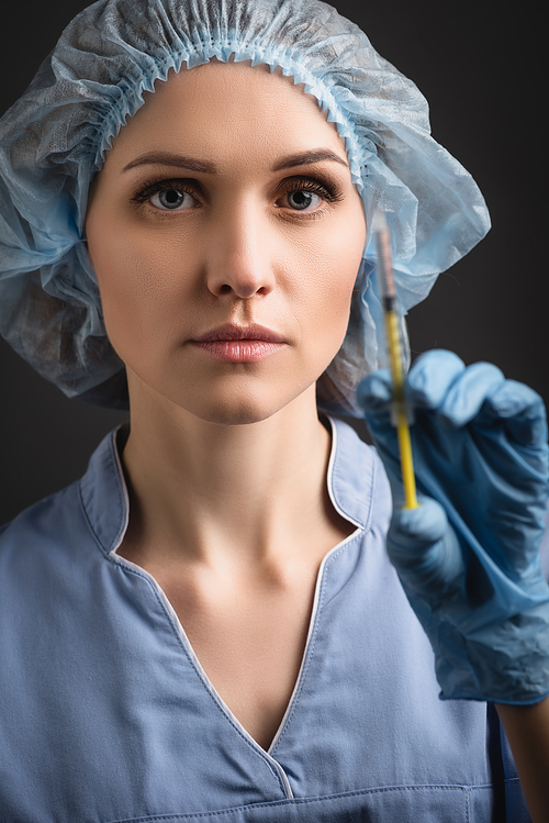 nurse in medical cap and uniform holding syringe with vaccine on blurred foreground isolated on dark grey