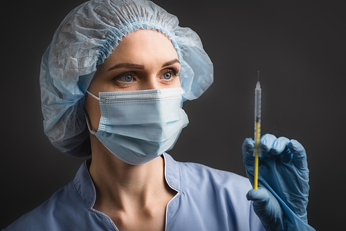 nurse in medical mask holding syringe with vaccine on blurred foreground isolated on dark grey