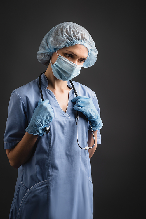 nurse in medical mask and latex gloves adjusting stethoscope isolated on dark grey