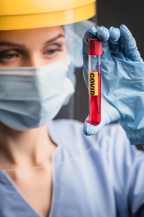test tube with covid lettering on hand of nurse in medical mask and face shield on blurred background