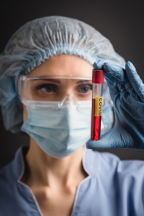 test tube with covid lettering on hand of nurse in medical cap, mask and goggles on blurred background isolated on dark grey