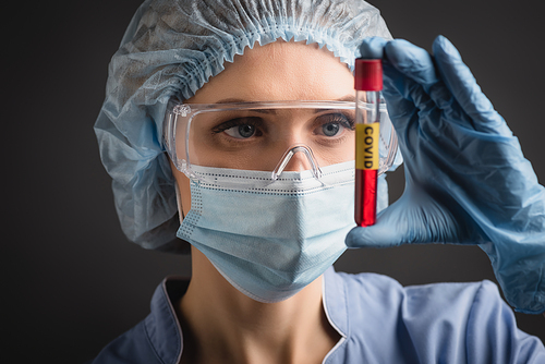 nurse in medical cap, mask and goggles holding test tube with covid lettering on blurred foreground isolated on dark grey