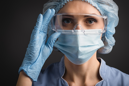 nurse in blue medical cap and mask adjusting goggles while  isolated on dark grey