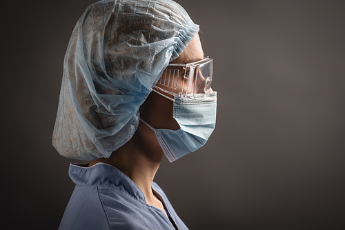 side view of nurse in medical cap, mask and goggles looking away isolated on dark grey