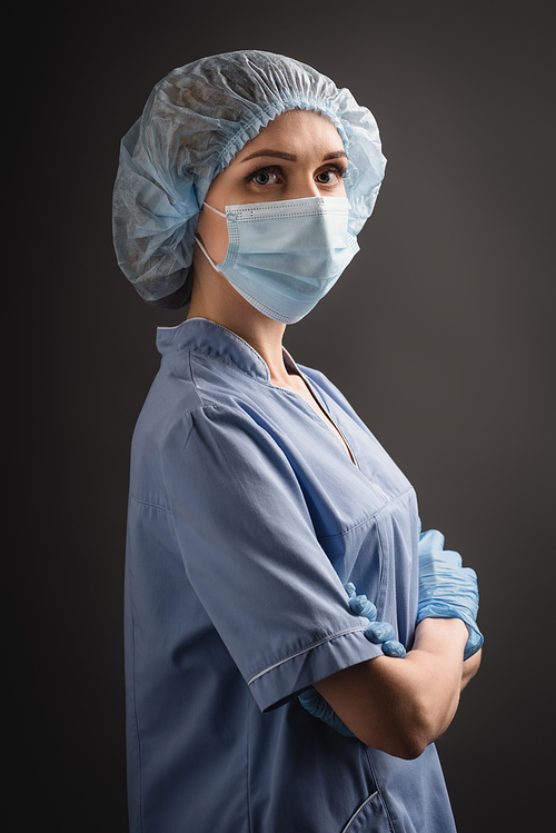 nurse in medical cap and mask standing with crossed arms and  isolated on dark grey