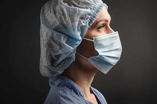 side view of nurse in medical cap and mask looking away isolated on dark grey
