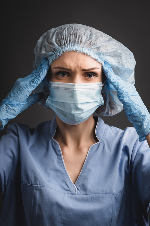 nurse in medical cap and mask suffering from headache isolated on dark grey