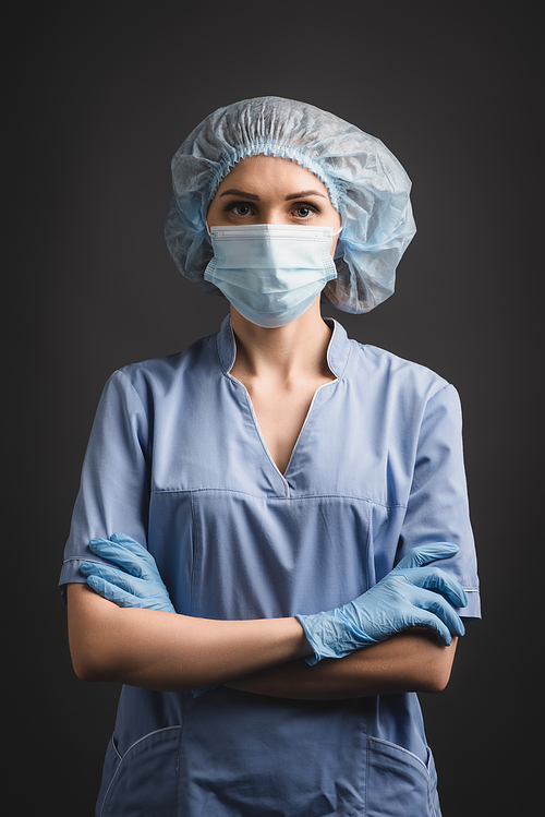 nurse in latex gloves, medical mask and cap standing with crossed arms isolated on dark grey