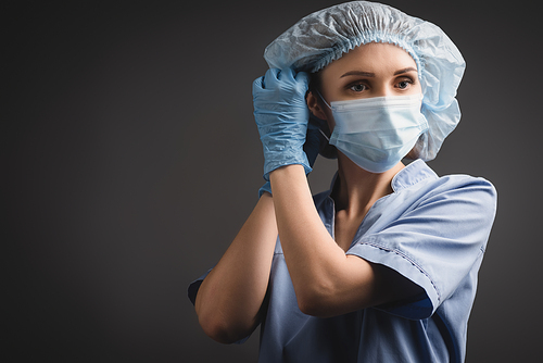 nurse in latex gloves adjusting medical cap isolated on dark grey