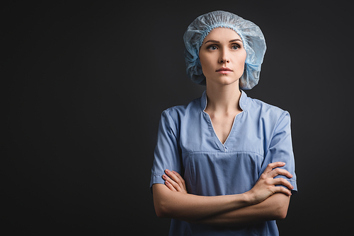 nurse in medical cap standing with crossed arms and looking away isolated on dark grey