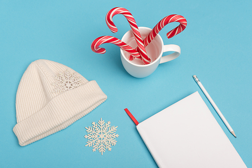 top view of beanie, candy canes and blank notebook on blue background