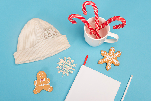 top view of gingerbread cookies, beanie, candy canes and blank notebook on blue background