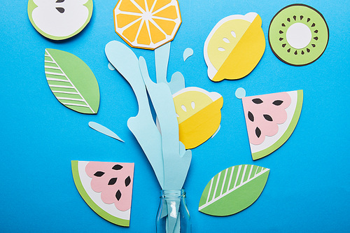 top view of paper cut water splash with fruits near glass bottle on blue background