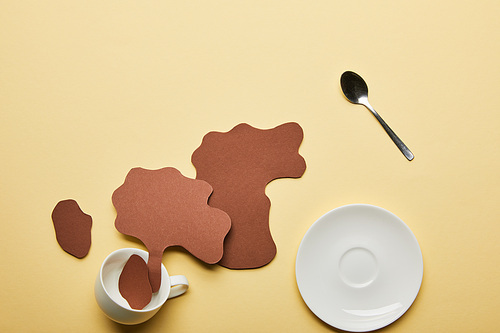 top view of paper cut coffee spills near white cup, saucer and spoon on beige background