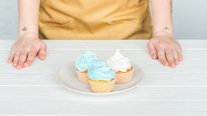 cropped view of woman near plate with delicious cupcakes on white table on grey