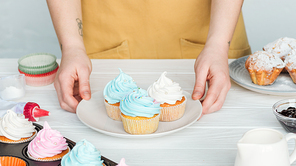 Cropped view of woman holding plate with cupcakes on table on grey