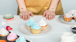 Cropped view of woman near table with plate with tasty cupcakes on grey
