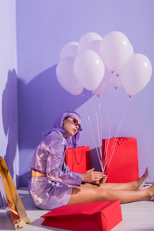 young woman dressed in doll style sitting with shopping bags and balloons on violet colorful background
