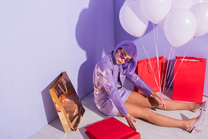 young woman dressed in doll style sitting with shopping bags and balloons on violet colorful background