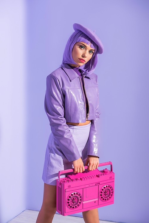young woman dressed in doll style posing with pink retro tape recorder on violet colorful background