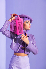 young woman dressed in doll style posing with pink retro tape recorder on violet colorful background