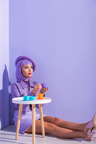 young woman dressed in doll style in beret posing with toy dishes on violet colorful background