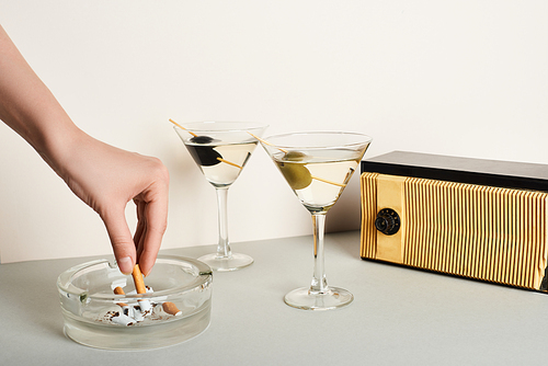 Cropped view of woman putting cigarette to ashtray beside cocktails and vintage radio on white background