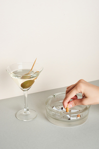 Cropped view of woman putting cigarette to ashtray with martini cocktail on grey surface