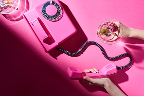 Cropped view of woman holding cocktail and telephone handset beside astray with cigarette butts on pink surface
