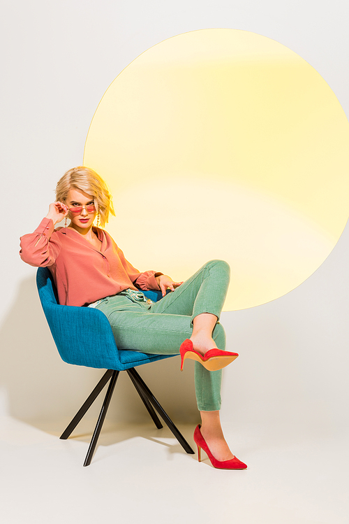 beautiful stylish young woman in colorful clothes sitting in armchair on white with yellow circle
