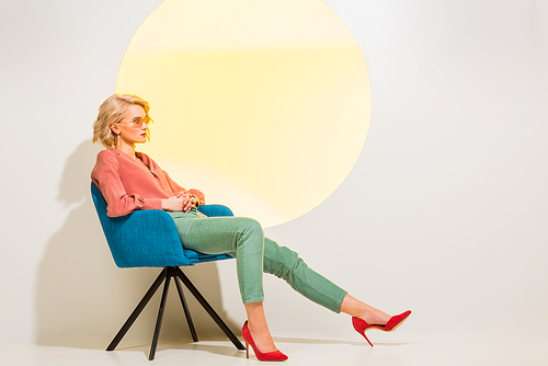 beautiful fashionable girl in colorful clothes sitting in armchair on white with yellow circle and copy space