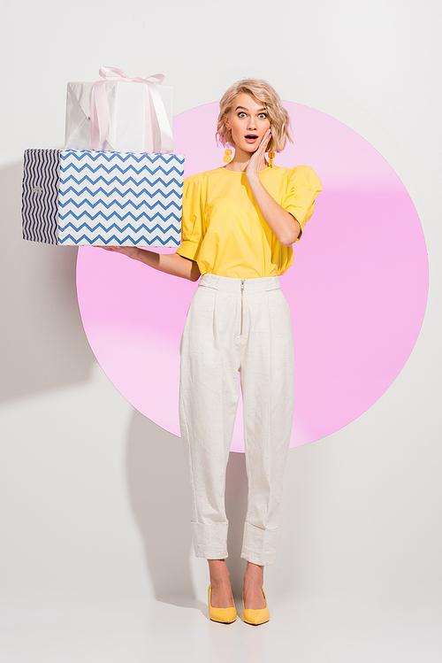 surprised fashionable girl gesturing and holding gift boxes on white with pink circle