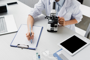 cropped view of scientist writing on clipboard near microscope and devices on desk