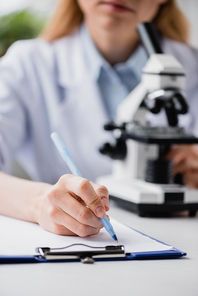 cropped view of scientist writing on clipboard near microscope on blurred background