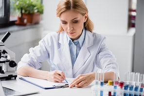 scientist writing on clipboard near microscope and test tubes on blurred background
