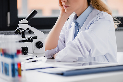 cropped view of scientist leaning on hand near microscope in lab
