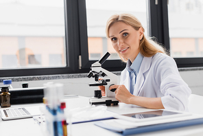 cheerful scientist in white coat looking at camera near microscope and digital tablet on desk