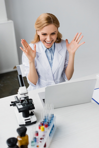 cheerful scientist in white coat looking at laptop near microscope on desk
