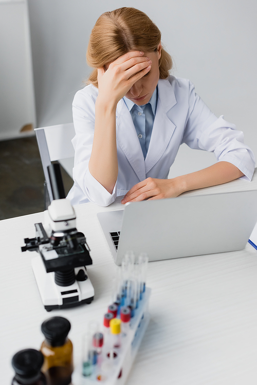 sad scientist  in white coat covering face near laptop and microscope on desk