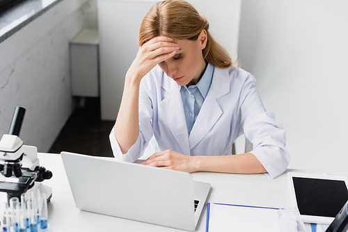 upset scientist in white coat covering face near laptop and microscope on desk