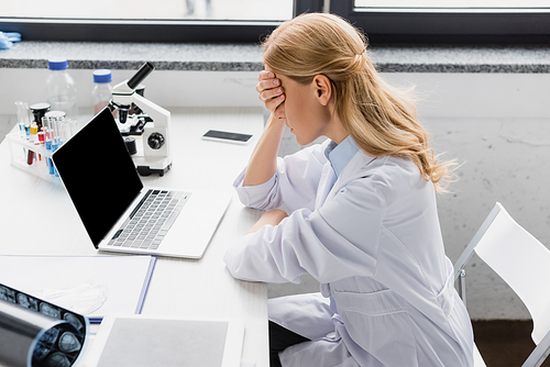 upset scientist in white coat covering face near laptop with blank screen and microscope on desk