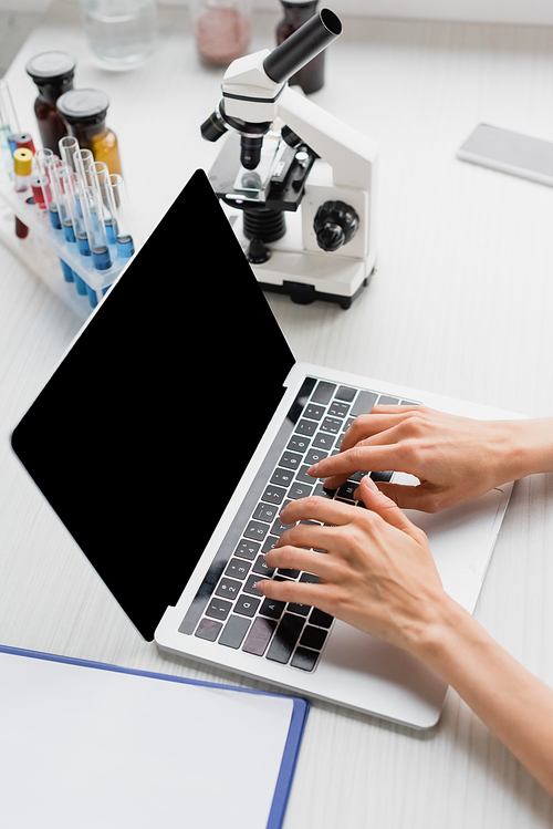 partial view of scientist typing on laptop with blank screen near microscope on desk