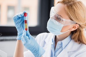 scientist in medical mask and goggles looking at test tube with covid lettering in lab
