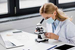 scientist in medical mask looking through microscope near gadgets with blank screen on desk