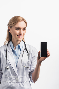 happy nurse in white coat smiling while holding smartphone with blank screen isolated on white