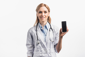 cheerful nurse in white coat smiling while holding smartphone with blank screen isolated on white