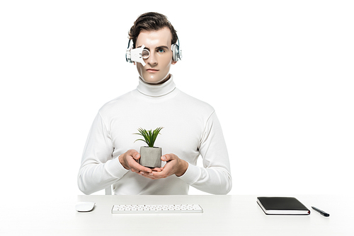 Cyborg in headphones and eye lens holding plant near computer mouse, keyboard and notebook isolated on white