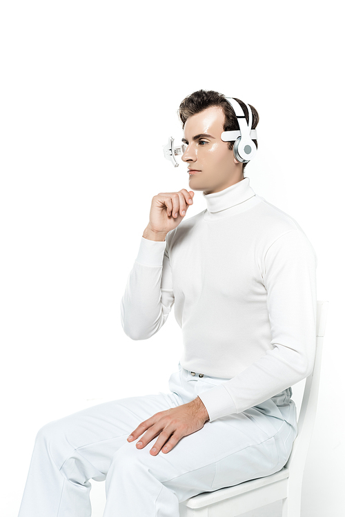 Cyborg man in eye lens and headphones with hand near chin sitting on chair isolated on white